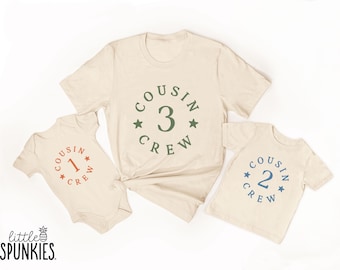 May include: A set of three cream-colored garments including one short-sleeve bodysuit and two t-shirts. Each features a circular graphic with the text "COUSIN CREW" and a number. The bodysuit has a rust-colored "1", one shirt has a blue "2", and one has a green "3".