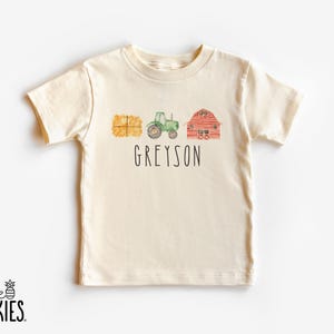 May include: A cream-colored short-sleeve t-shirt featuring a watercolor-style graphic of a yellow hay bale, a green tractor, and a red barn. Below the illustrations, the name GREYSON is printed in a simple, black, uppercase sans-serif font.