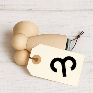 Könnte beinhalten: Ein Holz-Flaschenverschluss mit hellbraunem Finish. Ein cremefarbenes Etikett ist mit einer braunen Schnur befestigt und zeigt das schwarze Sternzeichen Widder. Der Verschluss ist für eine Flasche konzipiert.