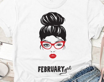 Puede incluir: Camiseta blanca con una ilustración en blanco y negro de una mujer con un moño en el pelo, gafas rojas y pintalabios rojo. El texto "FEBRUARY girl" está impreso debajo de la ilustración.