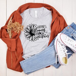 May include: A gray t-shirt with a black sunflower graphic and the text "Happiness blooms from within". The shirt is laid out on a white wooden surface with a brown jacket, blue jeans, and white sneakers.