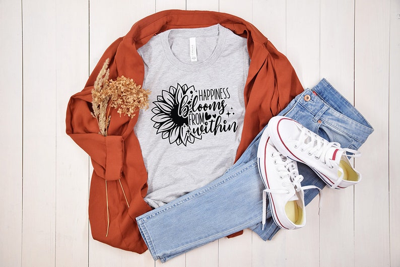 May include: A gray t-shirt with a black sunflower graphic and the text "Happiness blooms from within". The shirt is laid out on a white wooden surface with a brown jacket, blue jeans, and white sneakers.