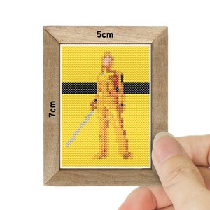 May include: A framed cross-stitch of a character from the movie "Kill Bill" wearing a yellow jumpsuit and holding a sword. The frame is 5 cm by 7 cm.