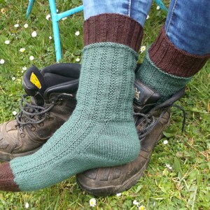 May include: A pair of green and brown knitted socks with a ribbed pattern. The socks are worn with brown leather boots and are sitting on a bed of green grass.