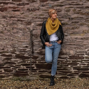 May include: A woman wearing a yellow knitted shawl, a black leather jacket, and blue jeans stands against a stone wall.