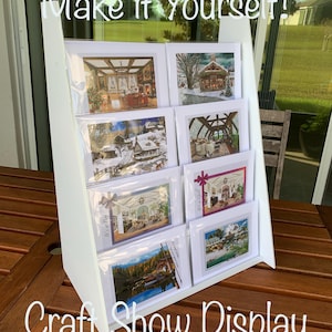 May include: White craft show display stand with multiple shelves, holding various greeting cards. The cards feature scenic images and are displayed on a wooden table. Text on the display reads "Make it Yourself!" and "Craft Show Display."