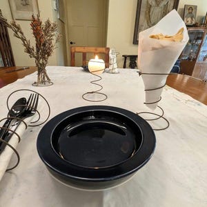 May include: A table setting with a navy blue plate, silverware wrapped in a white napkin, and a candle in a wire holder. A cone-shaped wire holder contains a paper cone filled with chips. Dried flowers are in a wire holder.