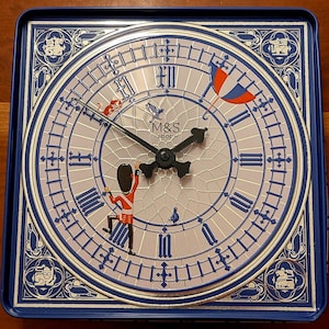 May include: A square blue metal tin featuring a clock face design with Roman numerals, black clock hands, and the M&S Food logo. The illustration includes a British guard figure, a small cat, a bird, and a red, white, and blue umbrella.