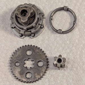 May include: An assortment of metal mechanical parts, including a circular assembly with multiple gears, a toothed ring, a large gear with three holes, and a small gear with a star-shaped center. The parts are arranged on a light-colored surface.