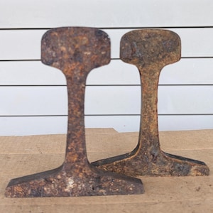 May include: Two upright sections of weathered, rusted steel railroad track. Each piece features a wide, flat base, a narrow vertical web, and a rounded, thick head at the top. The metal surfaces show significant oxidation with a dark brown texture.
