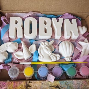 An A5 craft gift box featuring plaster letters personalised to the requested name for painting, alongside 6 themed shapes: 1 heart, 1 hot air balloon, 1 rainbow, 1 shooting star, 1 unicorn head and 1 unicorn horn with ears. Includes full size paintbrush, 6 colour themed paints and all bedded on a colour co-ordinated tissue paper dressing.