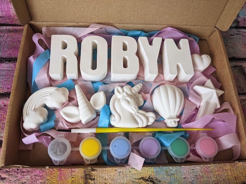 An A5 craft gift box featuring plaster letters personalised to the requested name for painting, alongside 6 themed shapes: 1 heart, 1 hot air balloon, 1 rainbow, 1 shooting star, 1 unicorn head and 1 unicorn horn with ears. Includes full size paintbrush, 6 colour themed paints and all bedded on a colour co-ordinated tissue paper dressing.