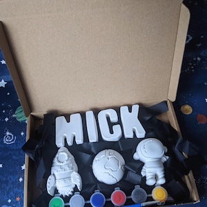 May include: A paint your own craft kit with a space theme. The kit includes a rocket, astronaut, globe, and the name "MICK" in large letters. The kit also includes a set of paint pots and a paintbrush.