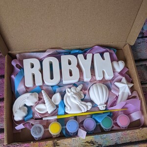 An A5 craft gift box featuring plaster letters personalised to the requested name for painting, alongside 6 themed shapes: 1 heart, 1 hot air balloon, 1 rainbow, 1 shooting star, 1 unicorn head and 1 unicorn horn with ears. Includes full size paintbrush, 6 colour themed paints and all bedded on a colour co-ordinated tissue paper dressing.