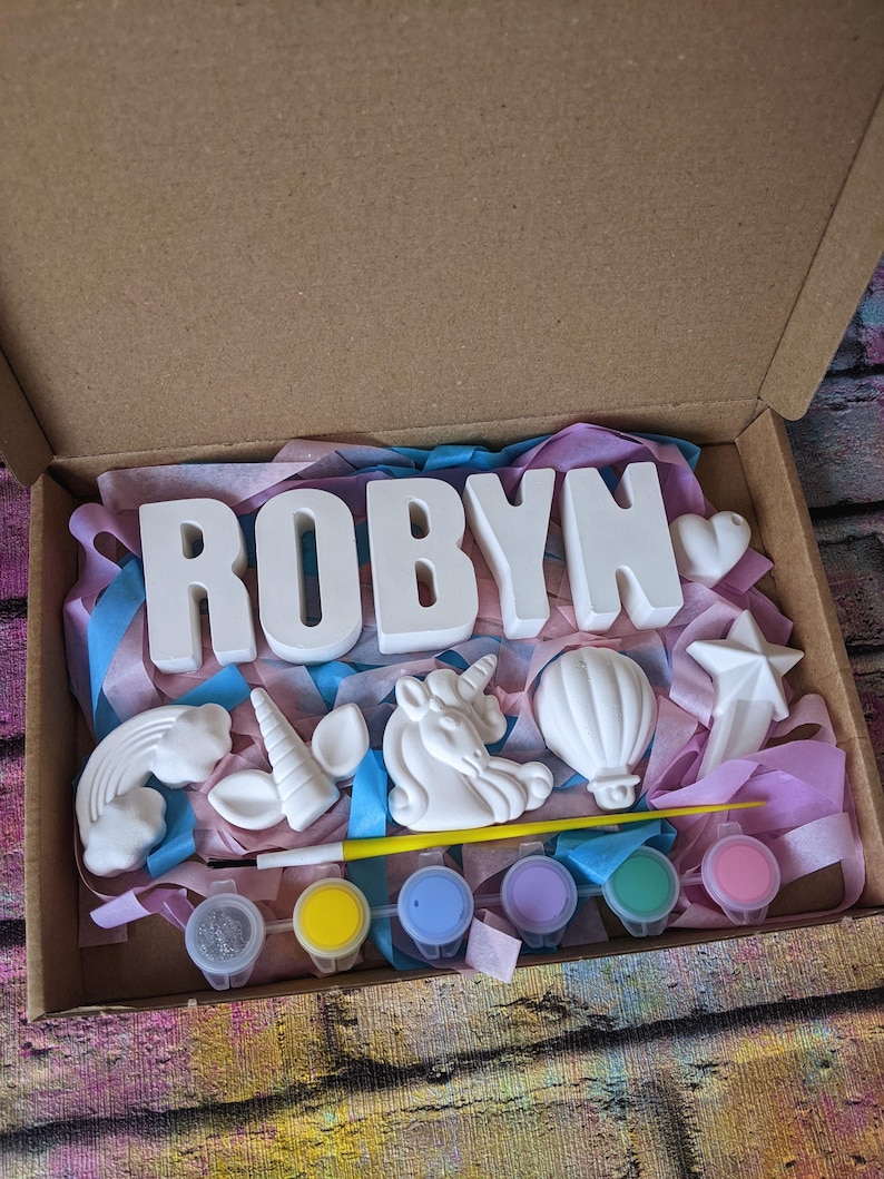 An A5 craft gift box featuring plaster letters personalised to the requested name for painting, alongside 6 themed shapes: 1 heart, 1 hot air balloon, 1 rainbow, 1 shooting star, 1 unicorn head and 1 unicorn horn with ears. Includes full size paintbrush, 6 colour themed paints and all bedded on a colour co-ordinated tissue paper dressing.