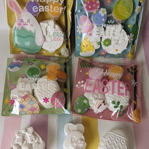 May include: A set of six Easter-themed paint-your-own ceramic ornaments. The ornaments include a bunny, an egg, a chick, and a basket. The ornaments are packaged in clear plastic bags with colourful backgrounds and the text "Happy Easter!"