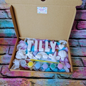An A5 craft gift box featuring plaster letters personalised to the requested name for painting, alongside 6 themed shapes: 1 heart, 1 hot air balloon, 1 rainbow, 1 shooting star, 1 unicorn head and 1 unicorn horn with ears. Includes full size paintbrush, 6 colour themed paints and all bedded on a colour co-ordinated tissue paper dressing.