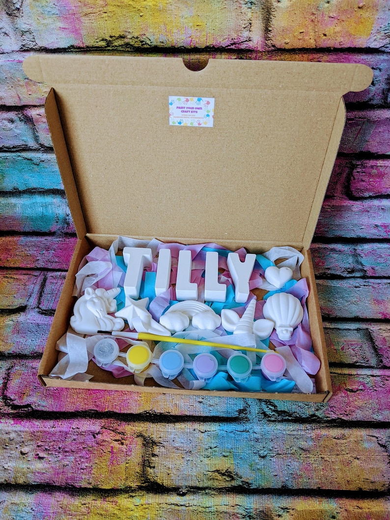 An A5 craft gift box featuring plaster letters personalised to the requested name for painting, alongside 6 themed shapes: 1 heart, 1 hot air balloon, 1 rainbow, 1 shooting star, 1 unicorn head and 1 unicorn horn with ears. Includes full size paintbrush, 6 colour themed paints and all bedded on a colour co-ordinated tissue paper dressing.
