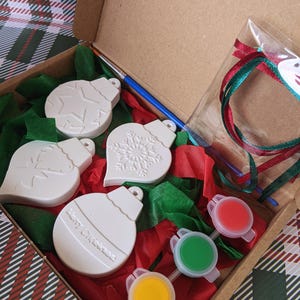 May include: A craft kit for making Christmas decorations. The kit includes four white ceramic baubles, three small pots of paint, a paintbrush, red and green tissue paper, and a red ribbon.