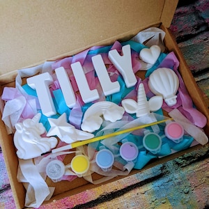 An A5 craft gift box featuring plaster letters personalised to the requested name for painting, alongside 6 themed shapes: 1 heart, 1 hot air balloon, 1 rainbow, 1 shooting star, 1 unicorn head and 1 unicorn horn with ears. Includes full size paintbrush, 6 colour themed paints and all bedded on a colour co-ordinated tissue paper dressing.