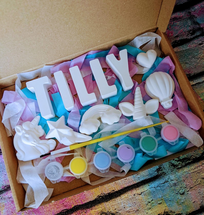 An A5 craft gift box featuring plaster letters personalised to the requested name for painting, alongside 6 themed shapes: 1 heart, 1 hot air balloon, 1 rainbow, 1 shooting star, 1 unicorn head and 1 unicorn horn with ears. Includes full size paintbrush, 6 colour themed paints and all bedded on a colour co-ordinated tissue paper dressing.