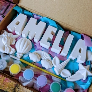 An A5 craft gift box featuring plaster letters personalised to the requested name for painting, alongside 6 themed shapes: 1 heart, 1 hot air balloon, 1 rainbow, 1 shooting star, 1 unicorn head and 1 unicorn horn with ears. Includes full size paintbrush, 6 colour themed paints and all bedded on a colour co-ordinated tissue paper dressing.