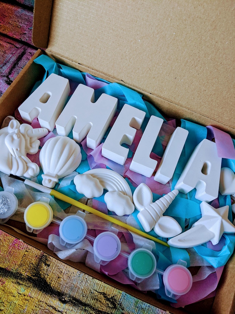 An A5 craft gift box featuring plaster letters personalised to the requested name for painting, alongside 6 themed shapes: 1 heart, 1 hot air balloon, 1 rainbow, 1 shooting star, 1 unicorn head and 1 unicorn horn with ears. Includes full size paintbrush, 6 colour themed paints and all bedded on a colour co-ordinated tissue paper dressing.