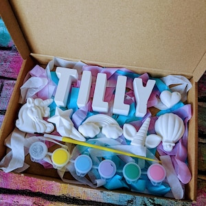 An A5 craft gift box featuring plaster letters personalised to the requested name for painting, alongside 6 themed shapes: 1 heart, 1 hot air balloon, 1 rainbow, 1 shooting star, 1 unicorn head and 1 unicorn horn with ears. Includes full size paintbrush, 6 colour themed paints and all bedded on a colour co-ordinated tissue paper dressing.
