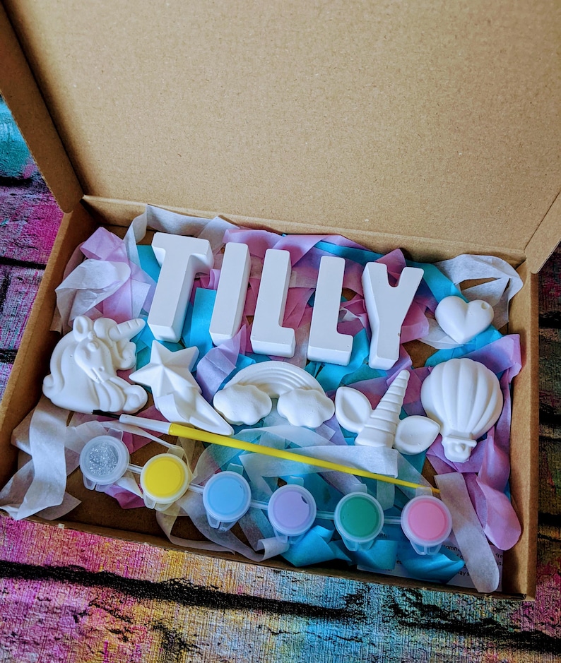 An A5 craft gift box featuring plaster letters personalised to the requested name for painting, alongside 6 themed shapes: 1 heart, 1 hot air balloon, 1 rainbow, 1 shooting star, 1 unicorn head and 1 unicorn horn with ears. Includes full size paintbrush, 6 colour themed paints and all bedded on a colour co-ordinated tissue paper dressing.
