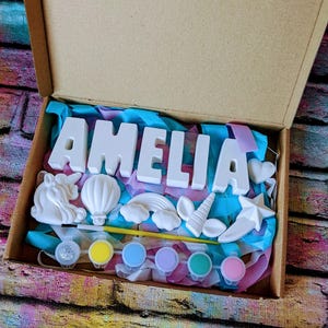 May include: A craft kit with white ceramic letters spelling "AMELIA" and unicorn, rainbow, star, and cloud shapes. The kit includes paint, a brush, and glitter.