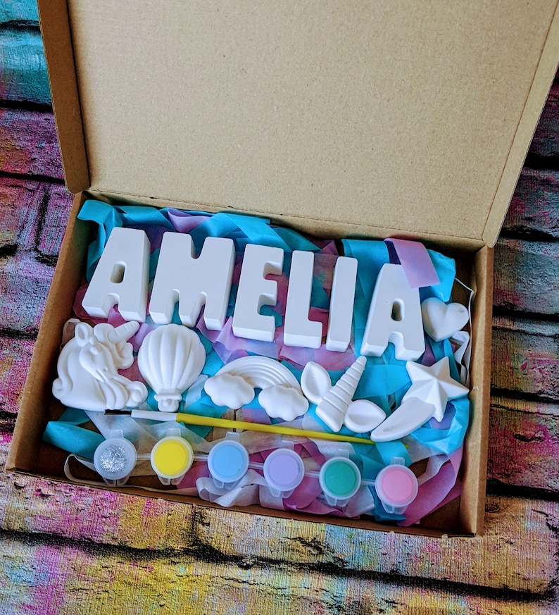 An A5 craft gift box featuring plaster letters personalised to the requested name for painting, alongside 6 themed shapes: 1 heart, 1 hot air balloon, 1 rainbow, 1 shooting star, 1 unicorn head and 1 unicorn horn with ears. Includes full size paintbrush, 6 colour themed paints and all bedded on a colour co-ordinated tissue paper dressing.