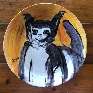 May include: A white ceramic plate with a hand-painted black and purple cat on a yellow background. The plate has the word "BYE" written on the bottom left side.