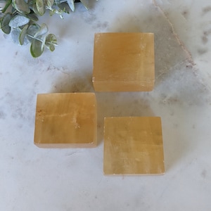 以下が含まれることがあります： 白い大理石の表面に置かれた、黄色のカルサイトの立方体が3つ。立方体は滑らかで半透明です。