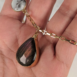 May include: A gold chain necklace with a teardrop-shaped iridescent labradorite gemstone pendant and a round white and black agate stone.