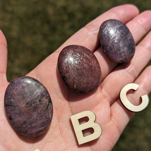 May include: Three smooth, polished purple and brown stones with a shimmery finish. The stones are arranged on a hand with wooden letters A, B, and C.