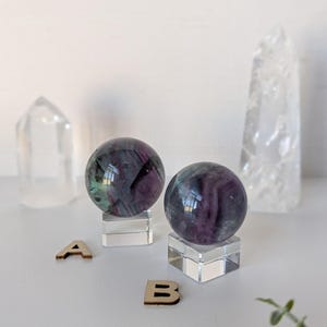 May include: Two fluorite spheres with purple and green hues sit on clear acrylic stands. Two clear quartz crystals and wooden letters 'A' and 'B' are also present. The spheres reflect light, creating a captivating display.