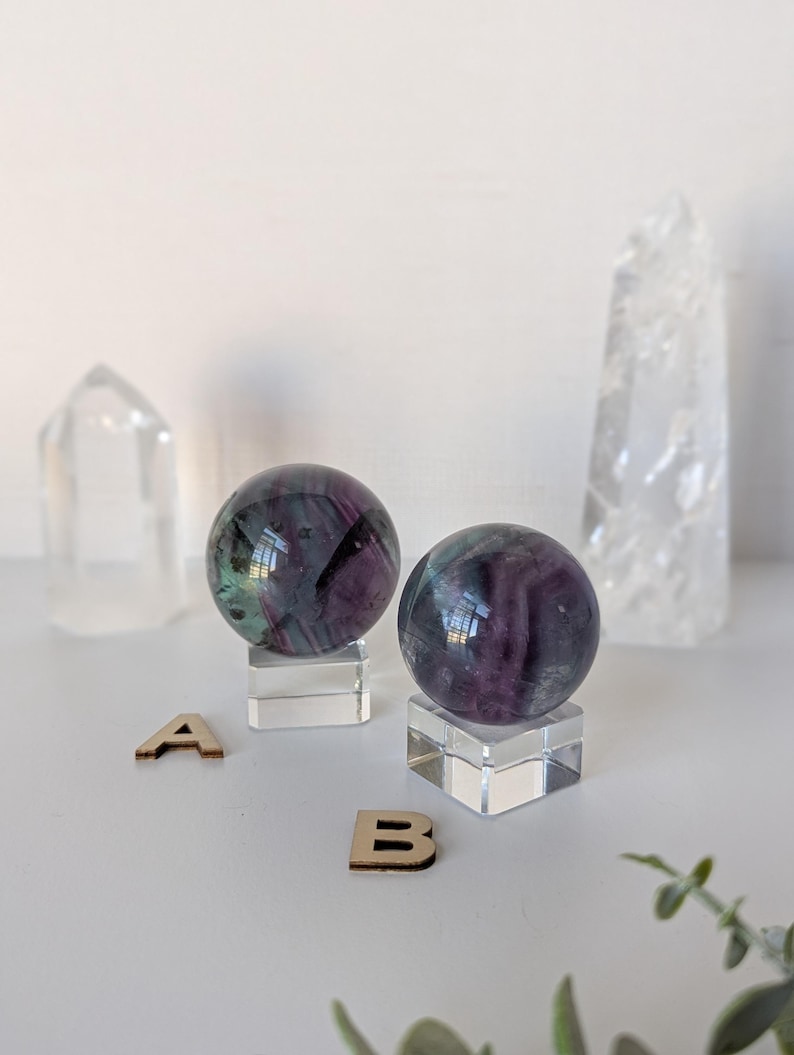 May include: Two fluorite spheres with purple and green hues sit on clear acrylic stands. Two clear quartz crystals and wooden letters 'A' and 'B' are also present. The spheres reflect light, creating a captivating display.