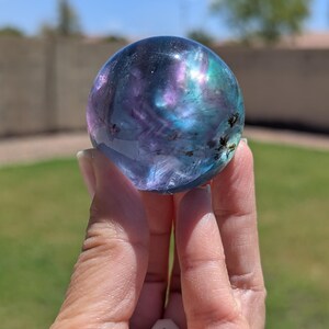 May include: A clear, spherical crystal ball with swirling patterns of purple, blue, and green. The crystal is held up against a blurred outdoor background, showcasing its iridescent colors and unique internal features.