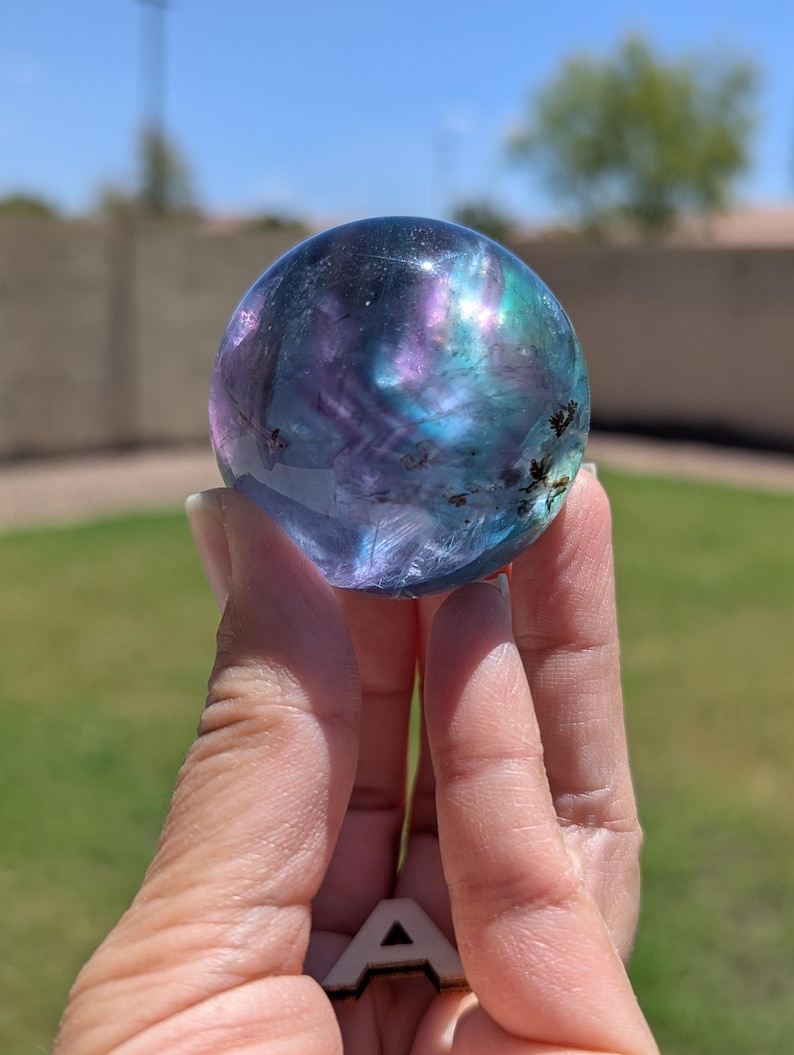 May include: A clear, spherical crystal ball with swirling patterns of purple, blue, and green. The crystal is held up against a blurred outdoor background, showcasing its iridescent colors and unique internal features.