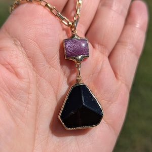 Puede incluir: Un collar de cadena de oro con un colgante que presenta una piedra preciosa negra facetada y una piedra preciosa púrpura más pequeña de forma cuadrada.