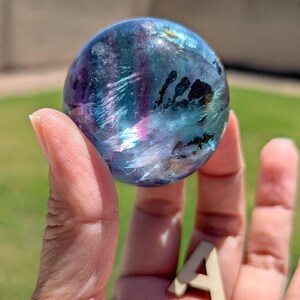 May include: A translucent, spherical crystal ball with swirling patterns of blue, purple, and hints of pink. The orb is held in a hand, with a small wooden letter "A" resting in the palm. The background is blurred, suggesting an outdoor setting.
