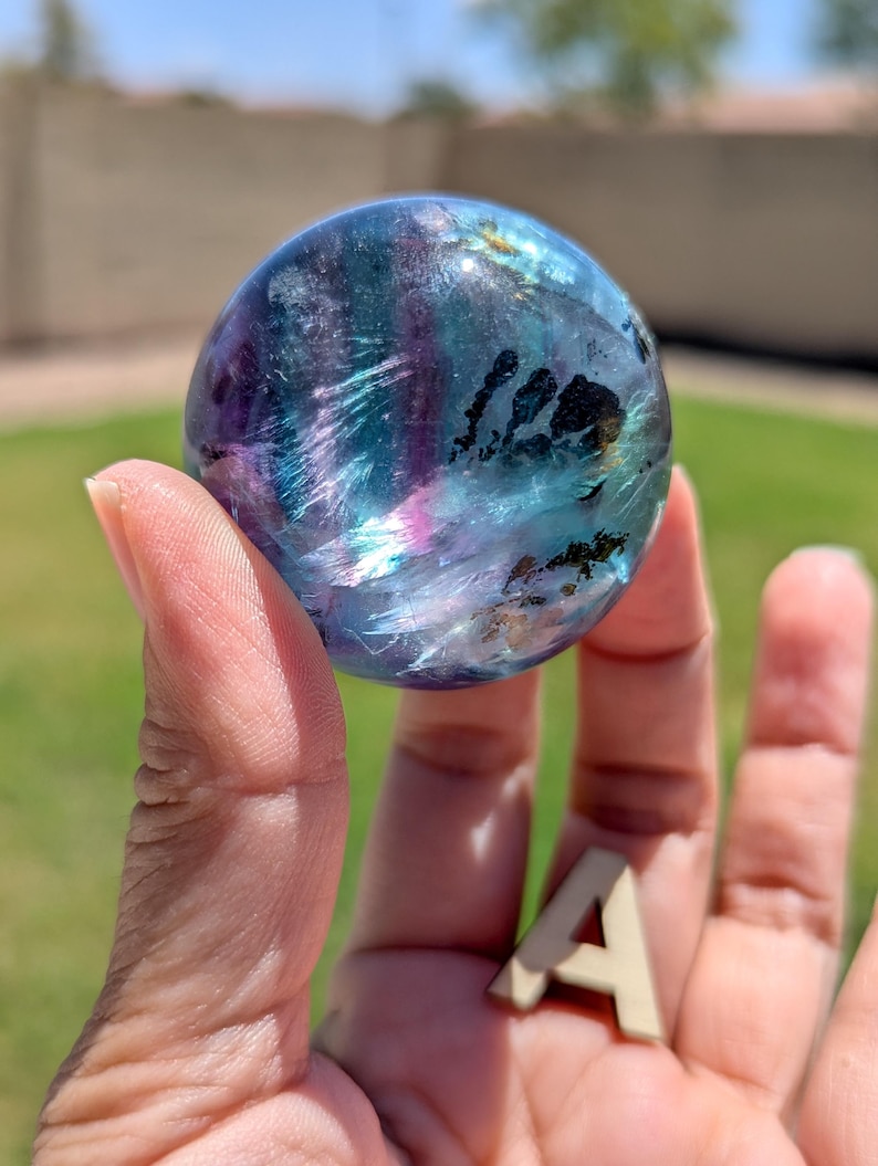 May include: A translucent, spherical crystal ball with swirling patterns of blue, purple, and hints of pink. The orb is held in a hand, with a small wooden letter "A" resting in the palm. The background is blurred, suggesting an outdoor setting.