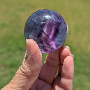 May include: A clear, spherical fluorite crystal ball with shades of purple and blue. The crystal ball is held in a hand against a blurred green background. The crystal ball is a decorative item.