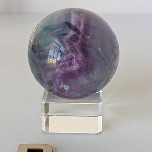 May include: A polished fluorite sphere, displaying a blend of purple, green, and clear hues. The crystal ball sits atop a clear, rectangular acrylic stand. The sphere's surface reflects light, showcasing its natural patterns and colors.