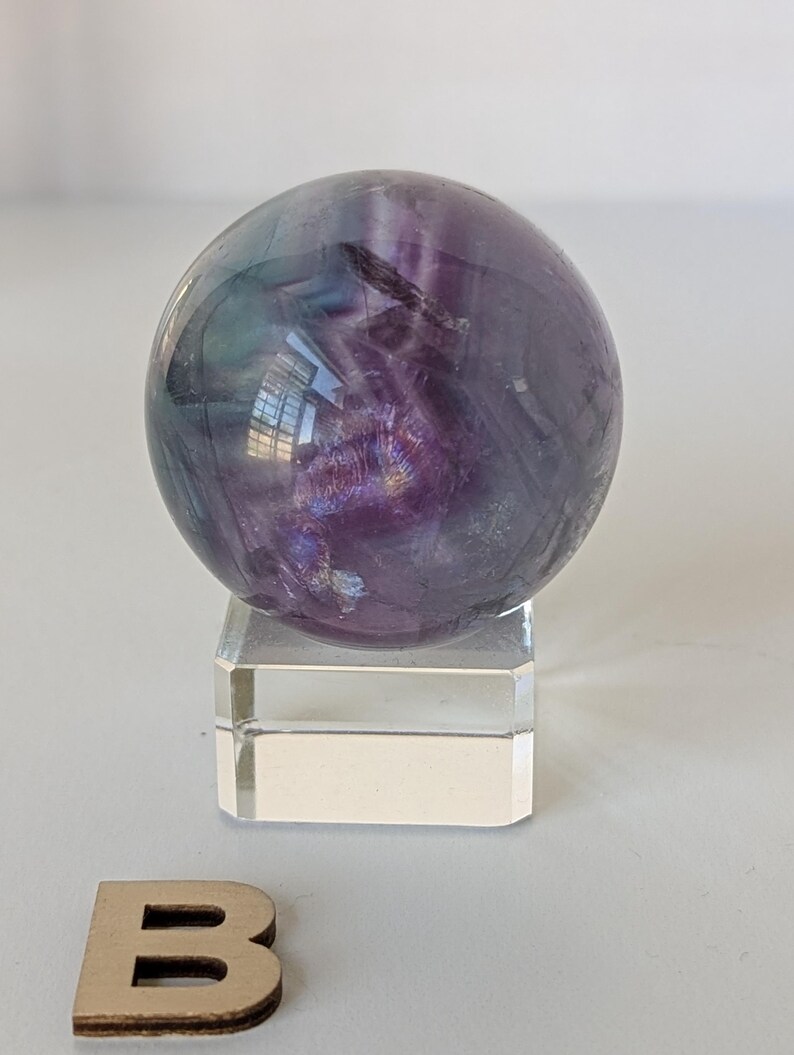 May include: A polished fluorite sphere, displaying a blend of purple, green, and clear hues. The crystal ball sits atop a clear, rectangular acrylic stand. The sphere's surface reflects light, showcasing its natural patterns and colors.