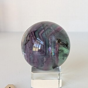 May include: A polished fluorite sphere, displaying bands of purple, green, and clear hues. The gemstone ball is mounted on a clear acrylic stand, showcasing its natural colors and patterns. The sphere is a decorative item.
