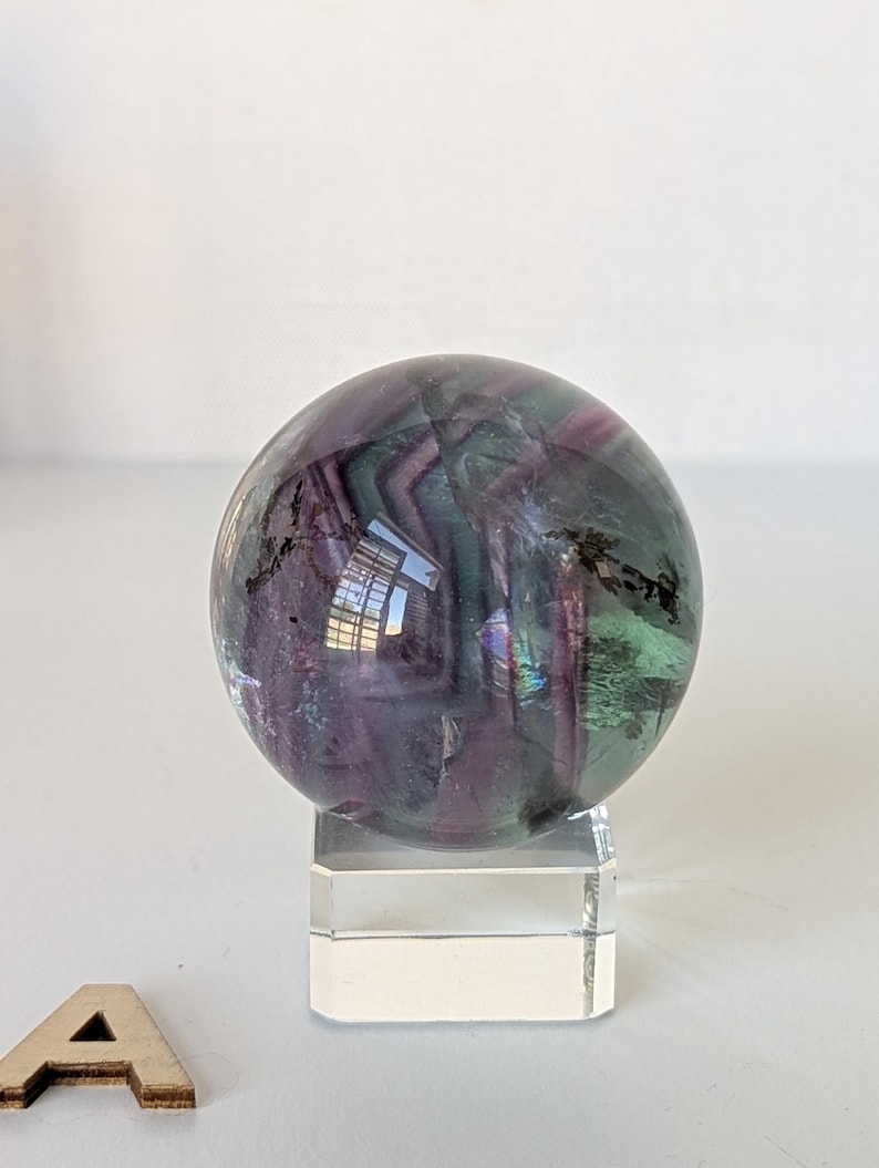 May include: A polished fluorite sphere, displaying bands of purple, green, and clear hues. The gemstone ball is mounted on a clear acrylic stand, showcasing its natural colors and patterns. The sphere is a decorative item.