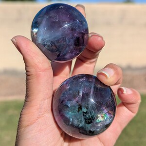 May include: Two polished fluorite spheres, held in a hand, showcasing a blend of purple, blue, and green hues. The spheres have a translucent quality, with internal patterns and a reflective surface. The background is blurred, suggesting an outdoor setting.