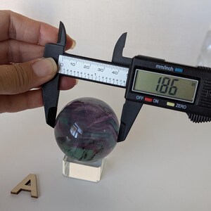 May include: A polished fluorite sphere, displaying swirls of purple and green, is measured with a digital caliper. The measurement reads 1.86 inches. The sphere rests on a clear acrylic stand, with a wooden letter "A" nearby.