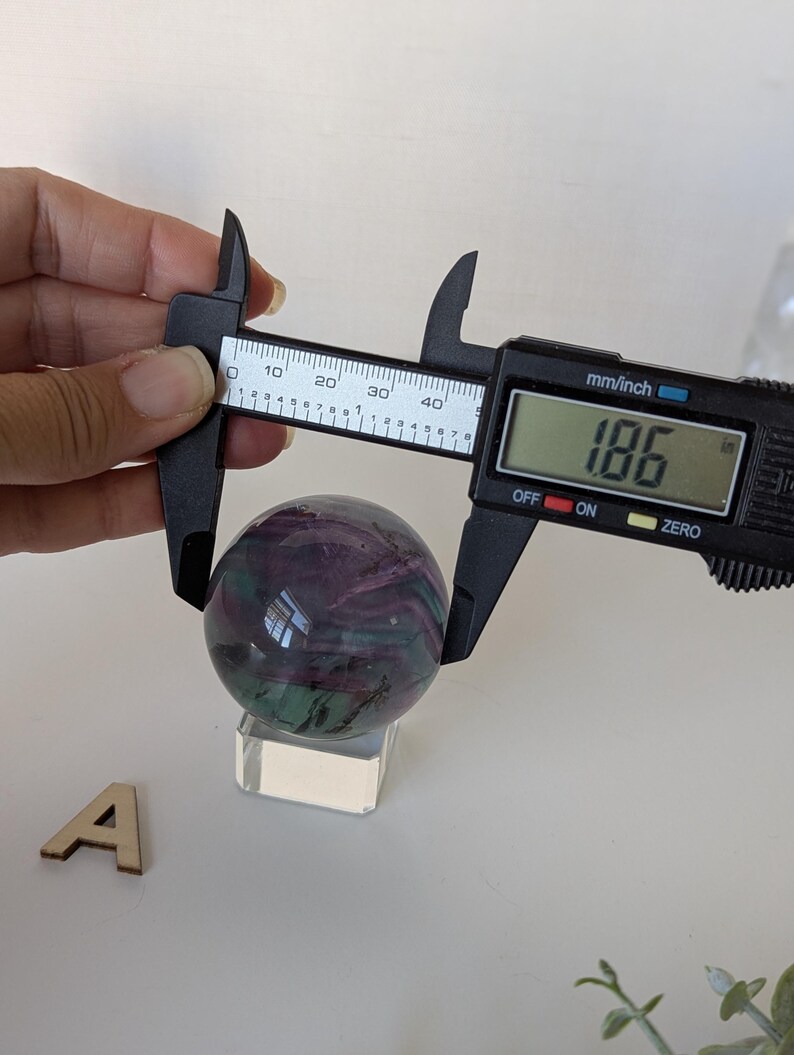 May include: A polished fluorite sphere, displaying swirls of purple and green, is measured with a digital caliper. The measurement reads 1.86 inches. The sphere rests on a clear acrylic stand, with a wooden letter "A" nearby.